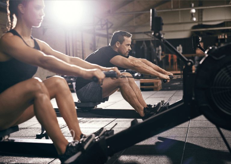 Man and woman on rowing machines in the gym, with the woman in the foreground; both have extended arms and bent legs, ready to push off.