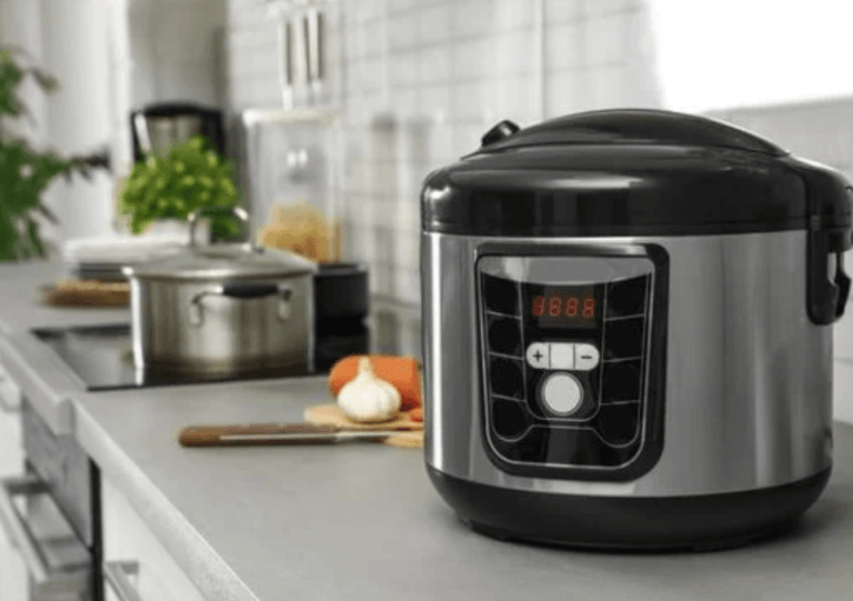 Slow cooker on a gray countertop with a stove, knife, garlic, and pan on an electric cooktop visible in the background.