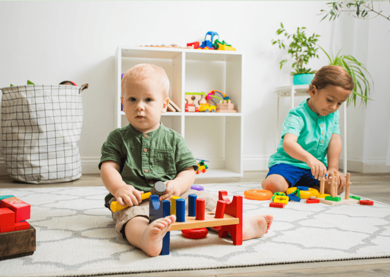 A pair of toddlers engaged with educational toys, including a hammering toy with wooden pieces. In the background, there’s a square Kallax shelf featuring four square compartments, creating a neat and inviting play space.