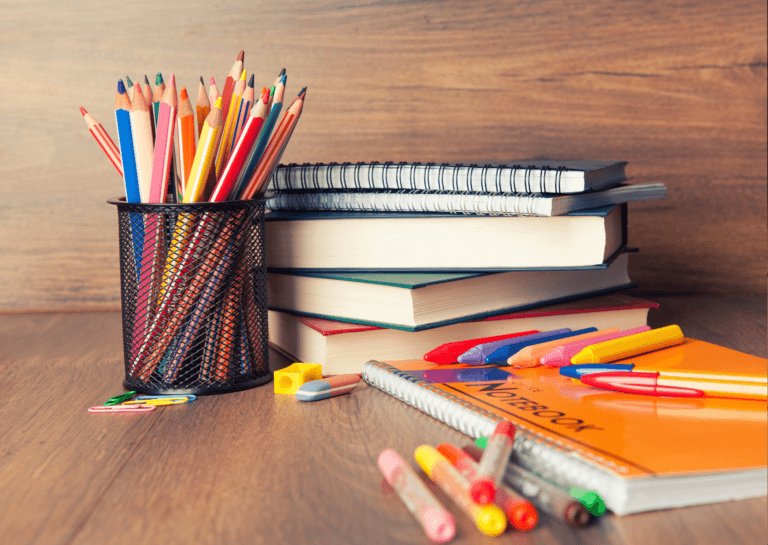 A collection of books stacked together, notebooks, and various colored pencils in a black metal cup holder. The items are placed on a wooden surface, creating a warm and inviting study atmosphere.