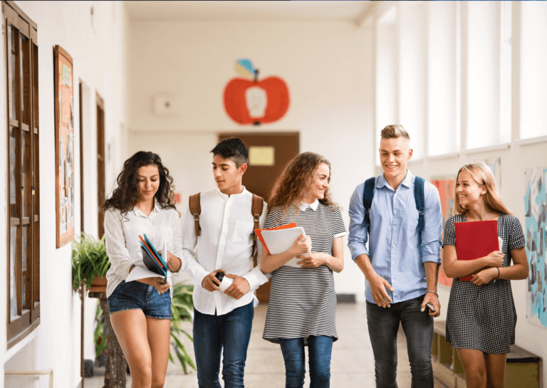 A group of five students, approximately 16 years old, walking down a school hallway and smiling. On the left is a girl, followed by a boy with a backpack, another girl in the center, another boy with a backpack, and a girl on the right. The girls each carry school papers in their hands.