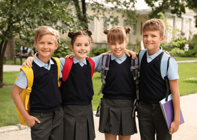 Two young boys on the outside and two young girls on the inside, standing with their arms crossed and smiling at the camera in school uniforms. They each carry a backpack, with a typical American school building several meters behind them.