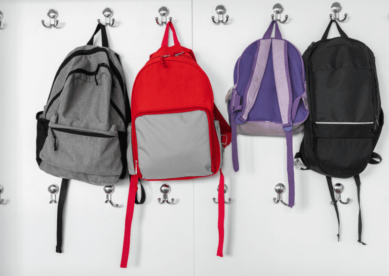 White wall featuring two rows of seven coat hooks, each with two hooks, displaying a total of four hanging backpacks in gray, red, purple, and black, arranged neatly side by side. The setup creates an organized and colorful storage solution.