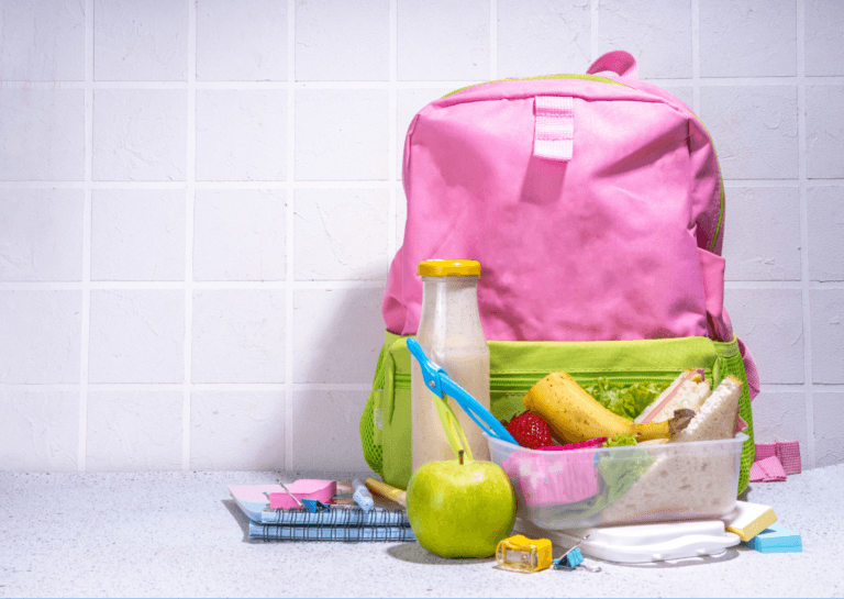 Schoolbag with a pink exterior and light green bottom standing on a countertop, accompanied by a transparent lunchbox containing sandwiches, half a banana, and a strawberry. In front of the bag are a green apple and a pencil sharpener, with small notepads beside the bag, all set against a kitchen wall made of 10x10 cm tiles.