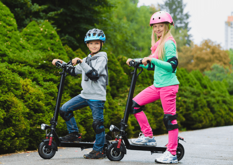 Young boy and girl, resembling siblings, standing still on electric scooters with their handlebars pointing to the left. The girl has blonde hair and wears a pink helmet, pink pants, and a green shirt, while her younger brother is in blue jeans and a gray sweater. Both are wearing safety knee and elbow pads.