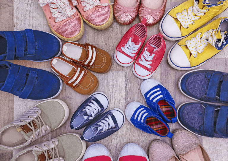 A variety of sneakers in different colors arranged in a circular formation with the toes facing each other. The outer ring features adult-sized shoes, while toddler shoes are placed in the center, all set against a light wooden floor, creating a playful and vibrant display.