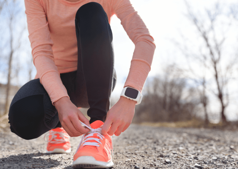 Girl crouched on the road tying the laces of her orange sports shoes, with one leg forward. She wears a white sports watch and is set against a backdrop of bare trees, creating an autumn or winter atmosphere on the rough asphalt.