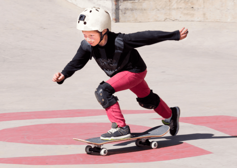 A young boy pushing off with one leg on a skateboard to gain speed, wearing a black sweater, white helmet, and red pants. He is on a light-colored floor with red accents, resembling a specific surface of a skate park.