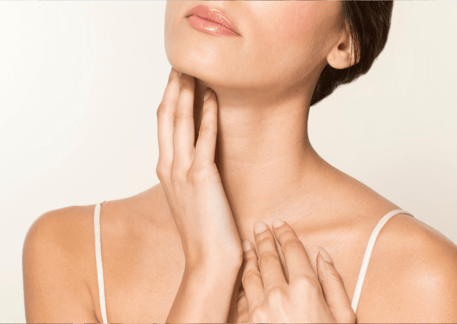 Young woman with brown hair and radiant skin, gently resting her hand on her face, showcasing her neck and the lower part of her face against a clean white background.