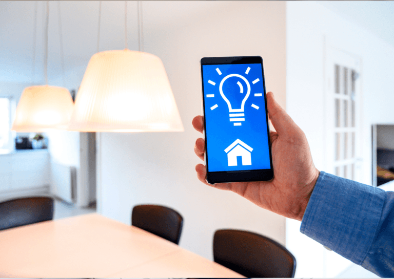 Person holding a smartphone displaying a virtual light bulb, controlling smart lighting above a dining table with hanging lamps turned on.