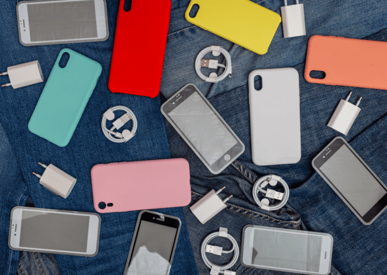 Variety of smartphones, phone cases, plugs, chargers, and charging cables in different colors, scattered haphazardly across various pairs of denim jeans.