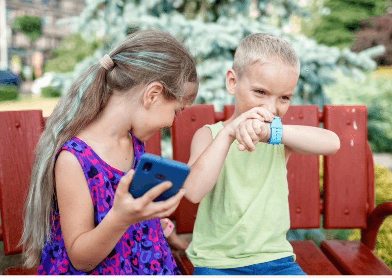 Young girl around 8 years old with long dark blonde hair gathered in a single tied ponytail, wearing a purple and red dress, sitting on a red bench with a boy around 5 years old in a sleeveless yellow shirt. The girl glances over her shoulder toward the boy, while he looks forward, speaking into his wrist with a watch.