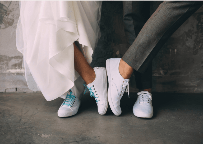 White new sneakers worn by both a man and a woman, with only their legs visible, as one foot touches the underside of the other shoe.