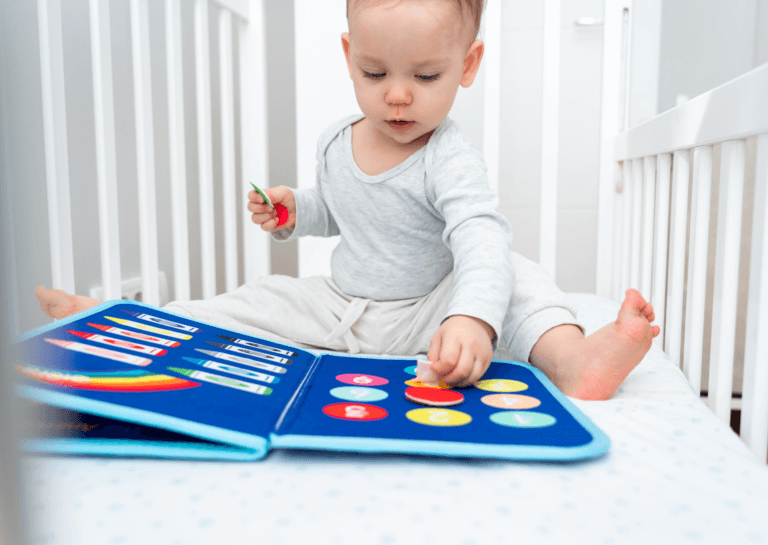 Toddler playing in a crib with a blue soft book, photographed from the front against a white background."