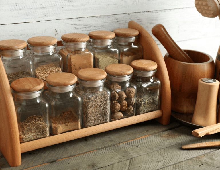 Wooden spice rack with 10 glass jars filled with various spices, each with a wooden lid, arranged in two staggered rows. A wooden salt shaker and spice mortar sit beside the rack.