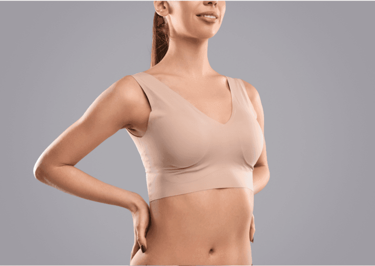 Thin young adult woman in a skin-toned sports bra, standing with hands on her hips, smiling; visible from just below her navel to her mouth, showcasing her bare abdomen.