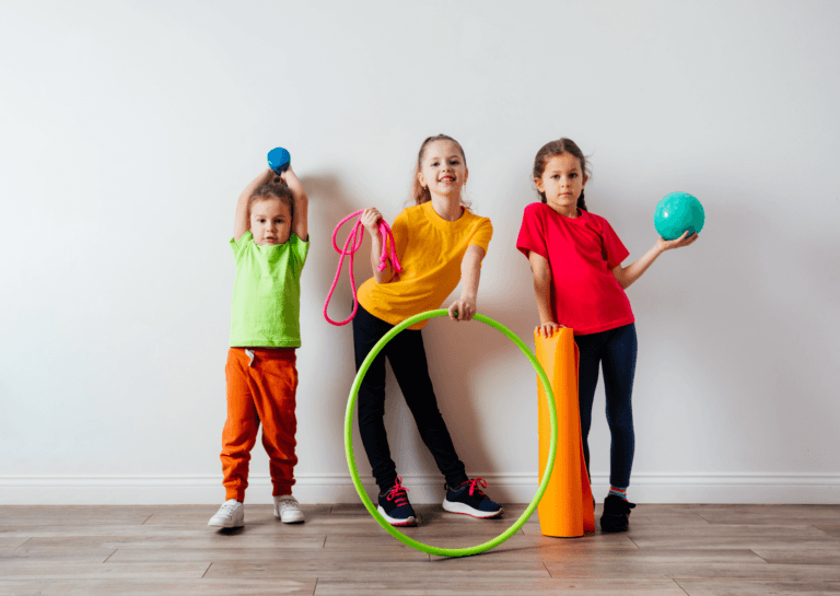 Three young girls, aged 4 and 7, standing side by side and looking at the camera while holding sports equipment like a hula hoop, rolled-up yoga mat, and a small fitness ball. They are dressed in colorful outfits, posing on a wooden floor in front of a white wall.