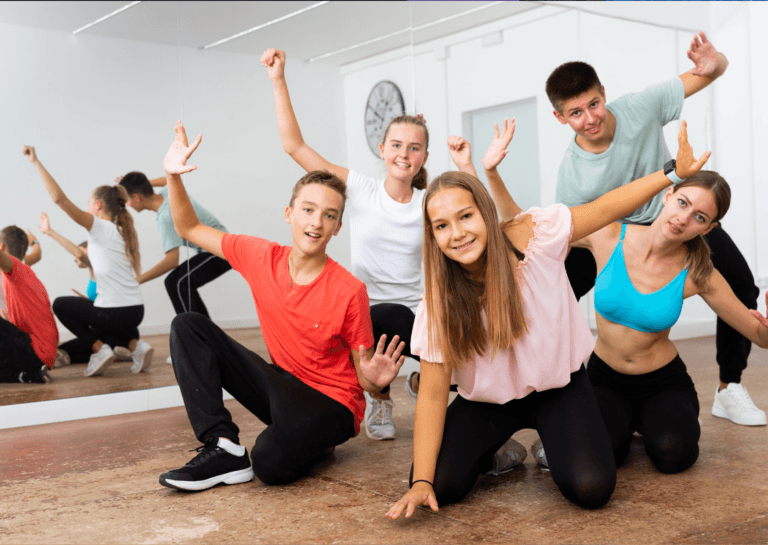 A group of five teens, three girls and two boys, crouching or bending forward with their hands in the air and smiling. They’re positioned in front of a large mirror wall in a sports hall, adding a sense of fun and energy to the scene.