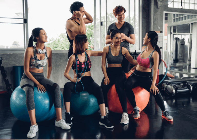 Two Asian men in sports clothing standing next to four young Asian girls in sportswear sitting on large fitness balls in a gym.