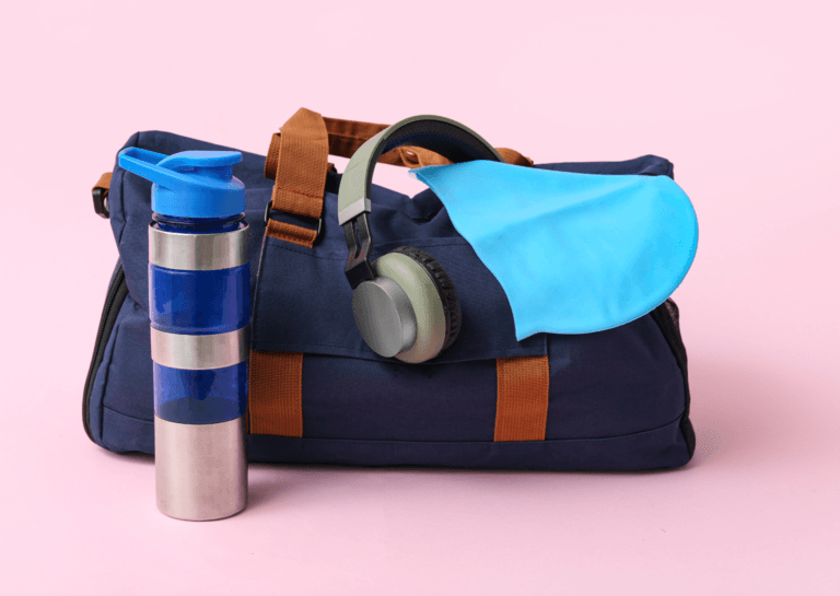 A blue sports bag with a brown handle, featuring a silver water bottle with a blue cap and two blue stripes resting in front of it. A pair of headphones is perched on the bag, all set against a pink background for a vibrant and stylish look.