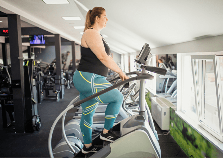 Plus-sized woman with blonde hair in a ponytail, wearing light green leggings and a sleeveless black top, using a stair climber in a gym; various fitness equipment and a small TV turned on are visible in the background, mounted on the ceiling.