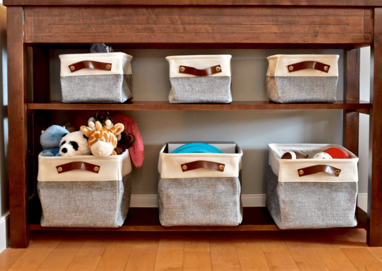 Storage solution for toddlers featuring six square bins with white tops and gray bottoms, organized in a brown cabinet with two shelves overflowing with toys.