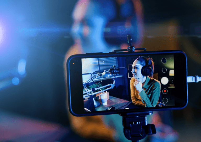 Smartphone in landscape mode placed in a phone holder, filming a woman with her hair tied back sitting at a desk in front of a microphone, with a closed white coffee cup in a dark environment featuring blinds in the background and a small desk lamp glowing behind her. The image on the smartphone shows the scene being recorded, faintly visible in the background.