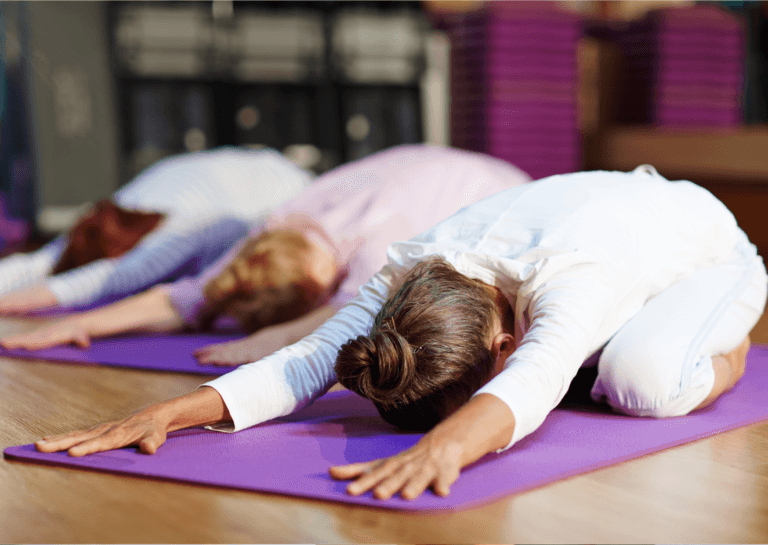 Three women in light outfits are stretching on purple mats, bending forward with their hands extended in front, positioned diagonally.