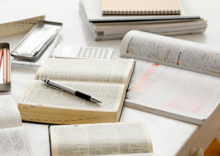 Open study book with a pen resting on its pages, surrounded by other books and notebooks on a white desk, creating a focused study setup.