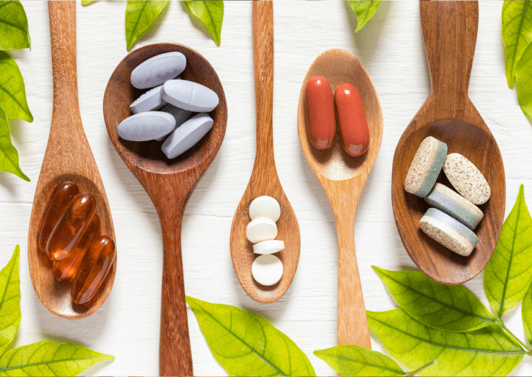 Various vitamin supplement capsules arranged alternately on wooden spoons, with a white background and green leaves at the sides, showcasing a natural and healthy theme.