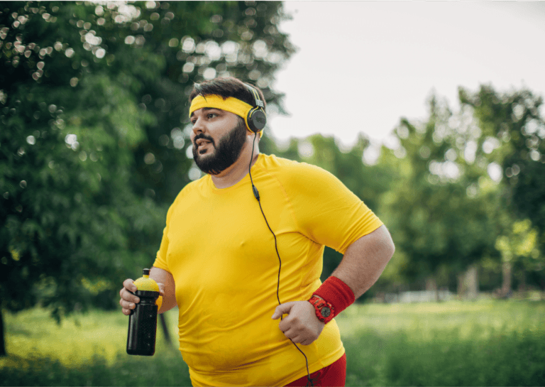 Plus-sized man wearing a yellow shirt and headband, with an orange arm sweatband, holding a black water bottle with a yellow cap, running with headphones visible, against a backdrop of lush greenery in a city park.