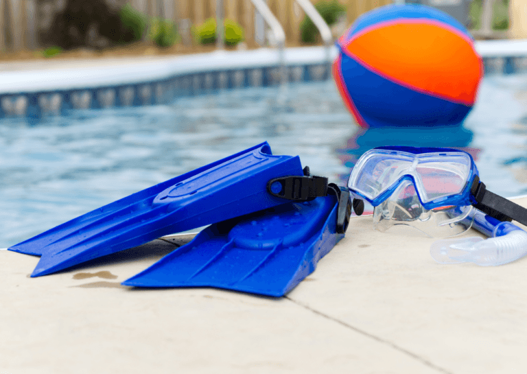 Blue swim fins lying over each other by the pool edge, with a blue and orange beach ball floating in the water and a diving mask set next to the fins.