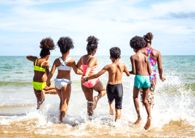 View from behind of six young African American children in swimwear holding hands as they joyfully enter the ocean."