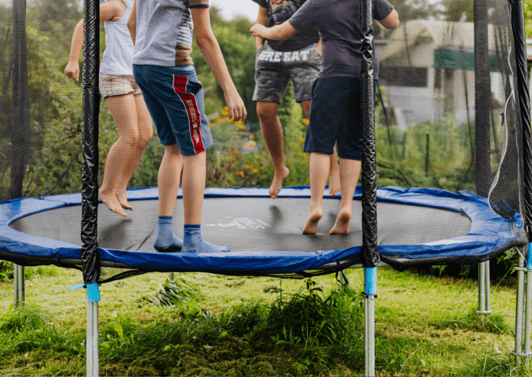 Four children jumping barefoot on a trampoline, surrounded by clear safety netting typical of freestanding trampolines, with their heads out of frame.