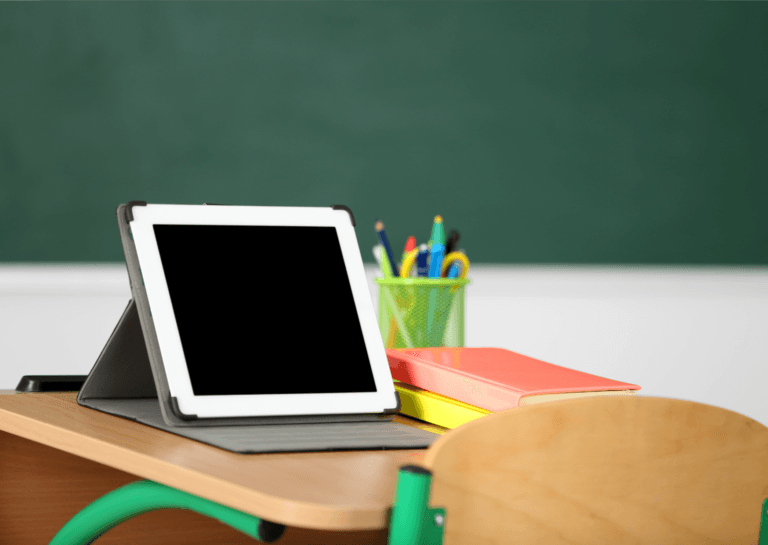 White tablet standing in a gray tablet stand on a school desk, with a green chalkboard in the background. The desk features a container of writing utensils and two notebooks, creating a vibrant classroom setting.