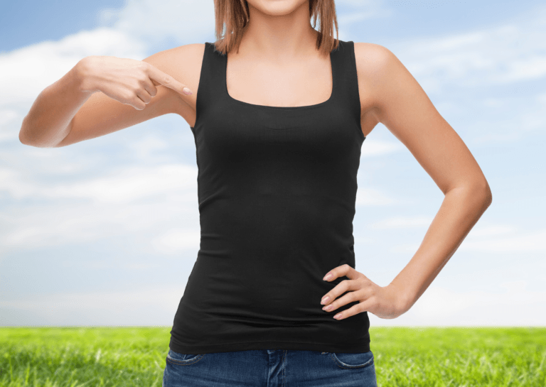 Upper body of a woman in a black tank top, pointing at her tank top with a horizon and green grass in the background.