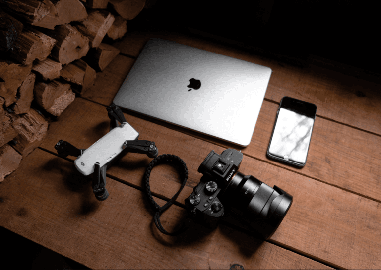 A closed silver-gray Apple laptop, a camera with a large lens, a white drone, and a smartphone are arranged on a wooden tabletop made of different planks. Chopped logs rest beside the laptop, adding a rustic touch to the setup.