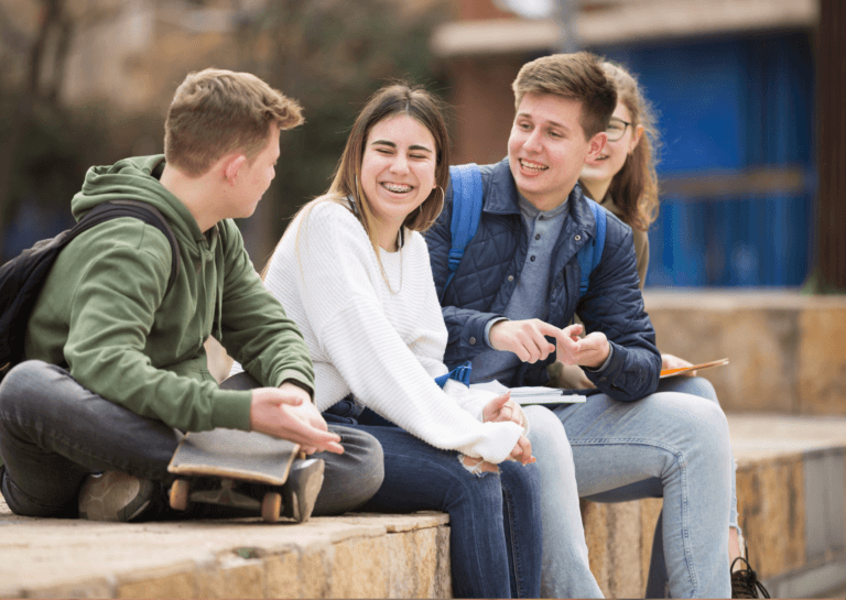 Two boys and two girls laughing while seated on a continuous rounded wooden log. Both boys are wearing backpacks, adding a casual vibe to the scene.