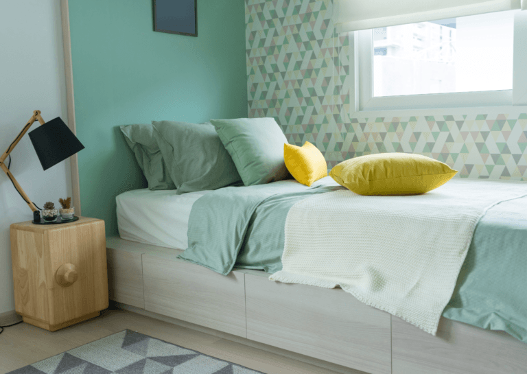 A small, tidy bedroom featuring a single bed adorned with three green pillows and two yellow ones, covered by a green duvet. A nightstand with a table lamp and a portion of a rug is visible, while a partially closed window with curtains above allows light to filter in.