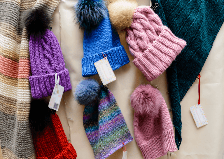 Six colorful hats hanging on a wall, arranged in two rows of three, with a string of lights draped between them (turned off) and a tag attached to each hat. The top row features a pair on the left, a blue one in the middle, and a pink knitted hat on the right. The bottom row includes a red hat on the left, a multicolored striped hat in the center, and a pink hat with a fluffy pom-pom on the right. The wall behind the hats is yellow, adding a cheerful touch to the display.