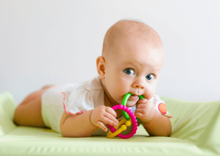 Baby lying on their stomach, chewing on teething toys while resting on a light green mattress.