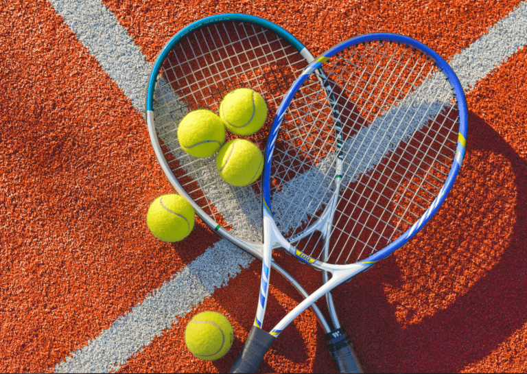 Two white and blue tennis rackets resting on the gravel at the intersection of the tennis court lines, with three tennis balls on one racket and two additional balls beside it.