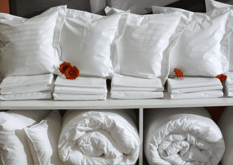 Toddler bedding sets with white pillows, folded duvet covers, and rolled blankets on a white cabinet, interspersed with a few loose red flowers.