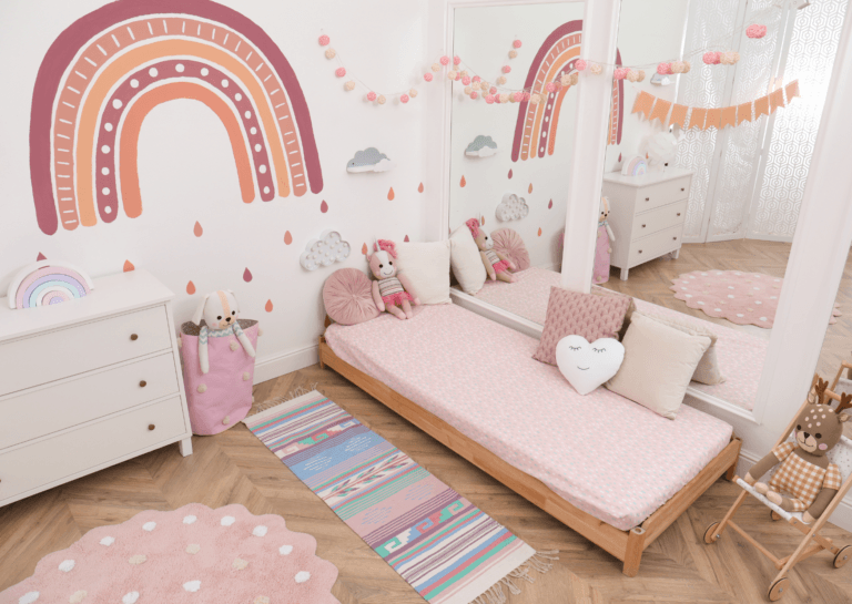 A charming toddler bedroom decorated in soft white and pink tones, featuring a rainbow design on the wall. The cozy atmosphere creates a delightful space for young girls to play and dream.