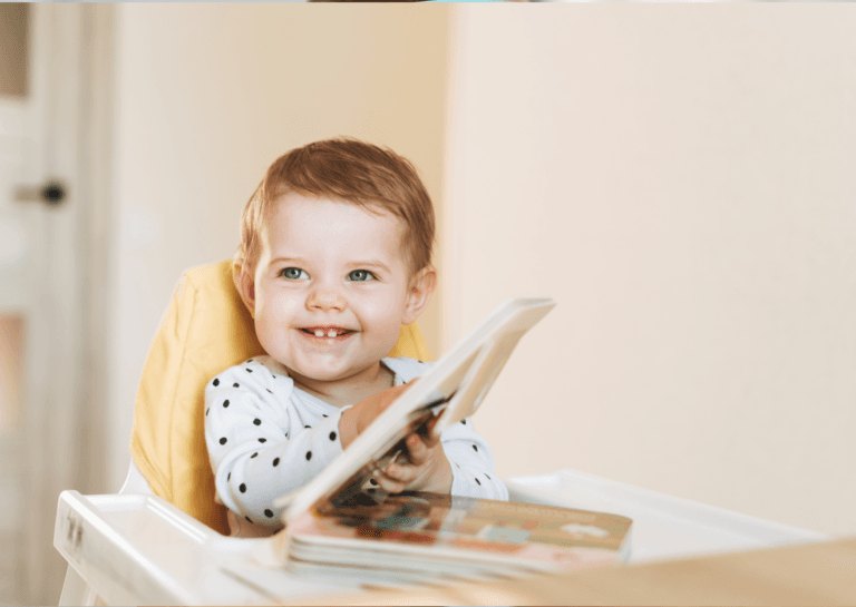 Toddler in a booster seat, intently browsing a book, with two prominent front teeth showing as they smile.