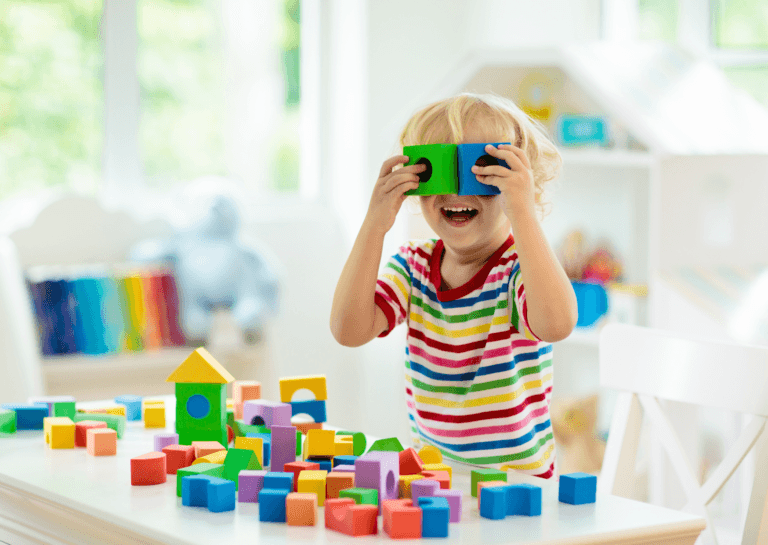 A cheerful blond boy holding two play blocks in front of his eyes, looking through them and laughing. His playful expression highlights the joy of learning through interactive toys.