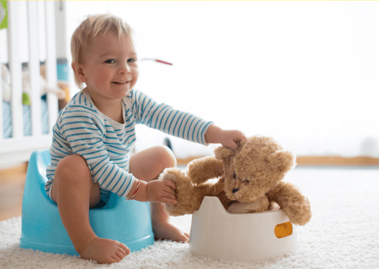 Toddler in a striped white and blue shirt sitting on a potty, placing a teddy bear on another potty in a playful scene.