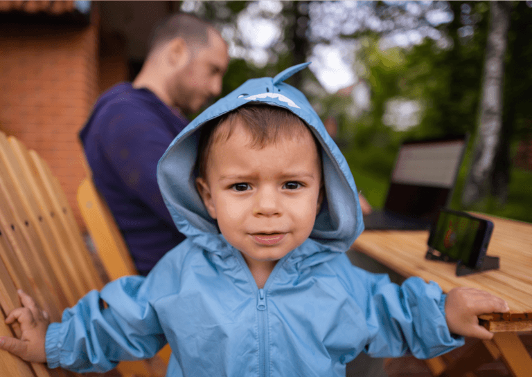 Toddler in a light blue rain jacket with a hood stands in front of a garden chair, looking at the camera, while father sits nearby at the garden table.