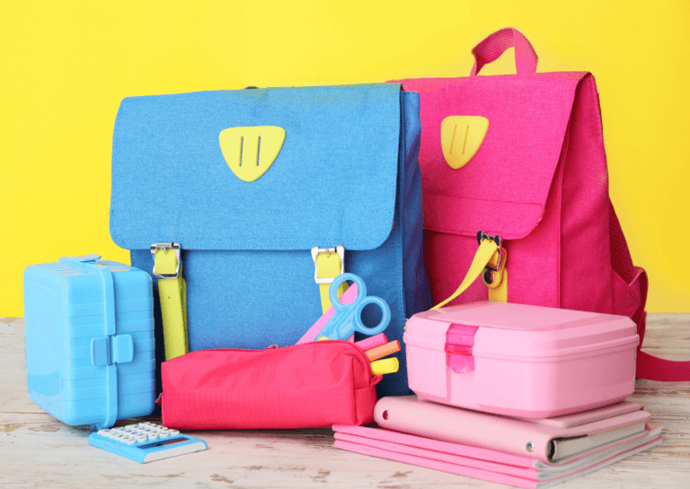 Colorful toddler backpacks and lunchboxes featuring a blue and red bag, light blue and pink lunchbox, red pencil case with markers, a blue calculator, and pink notebooks against a bright yellow background.