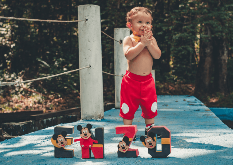 Toddler in red shorts and bare torso standing on a blue floor with building blocks spelling 'ENZO' in front of him.